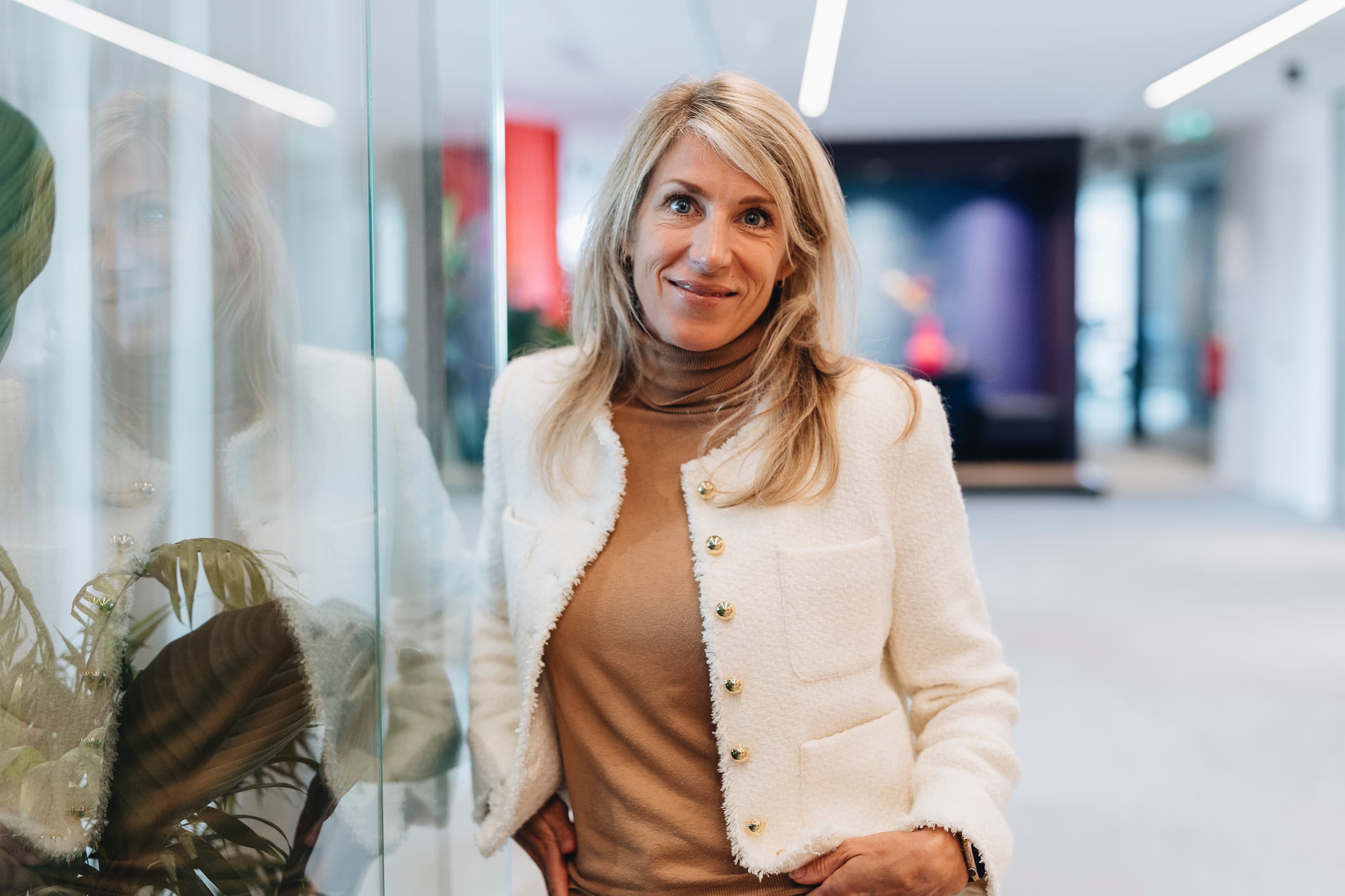 Vrouw in witte blazer en beige top in een moderne kantoorhal, met glazen wand en reflectie op de achtergrond