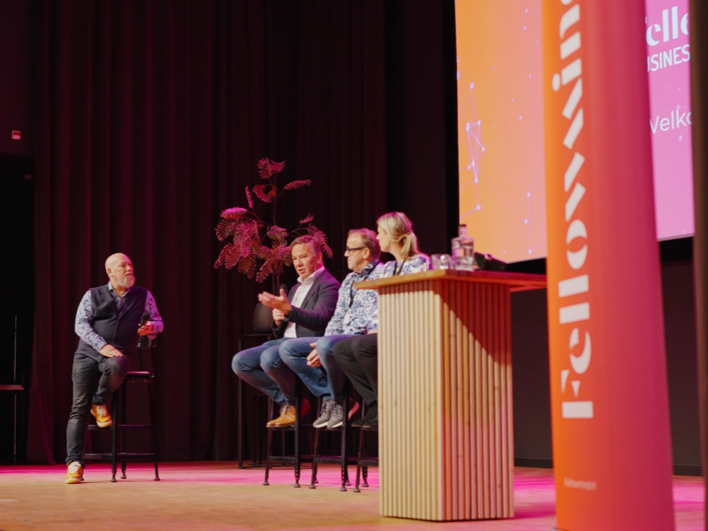 Paneldiscussie op een conferentiepodium met vier sprekers op barkrukken, microfoon en kleurrijke branding op de achtergrond
