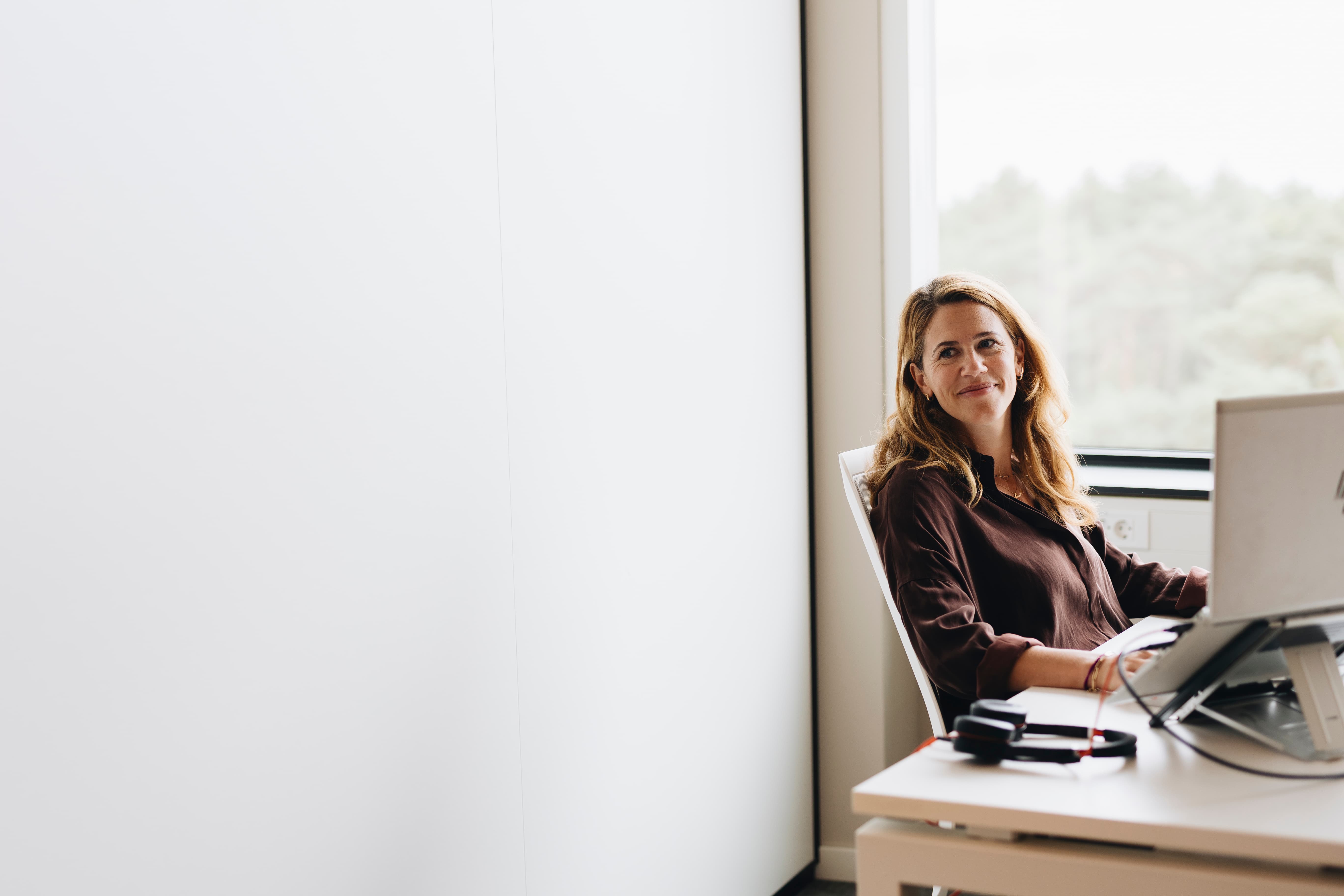 Vrouw met krullen leest een magazine bij een groot raam in een modern interieur met groene kamerplanten