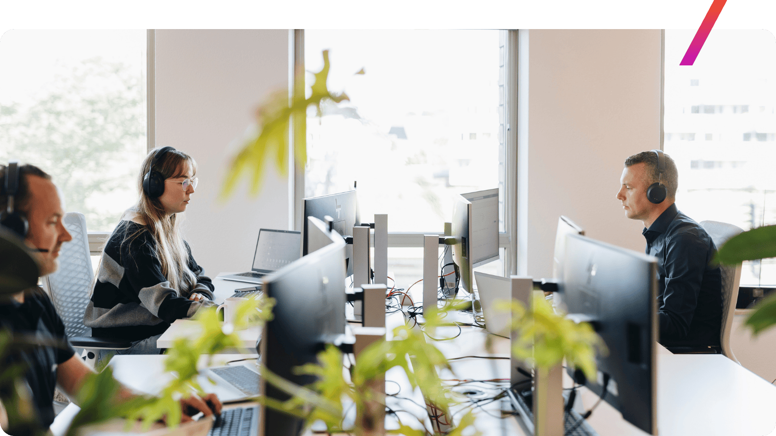 Medewerkers met koptelefoons werken achter computers in een modern kantoor, met veel daglicht en planten op de voorgrond