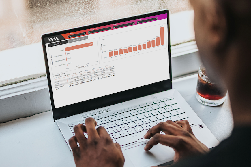 Person som arbetar på laptop med analysdashboard och stapeldiagram, bredvid ett glas dryck vid ett fönster