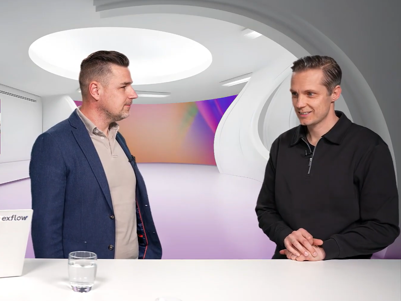 Twee mannen in een modern tv-studio-interieur, staand achter een witte tafel met glas water
