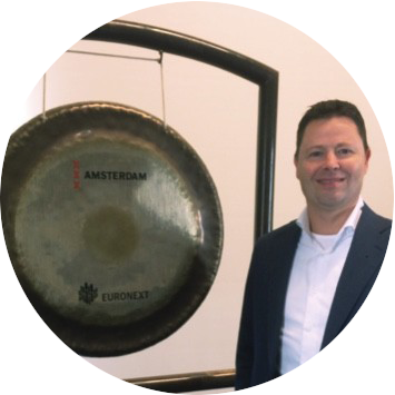 Een man in een donker pak staat naast een grote gong met Amsterdam- en Euronext-opdruk, tijdens een zakelijk evenement