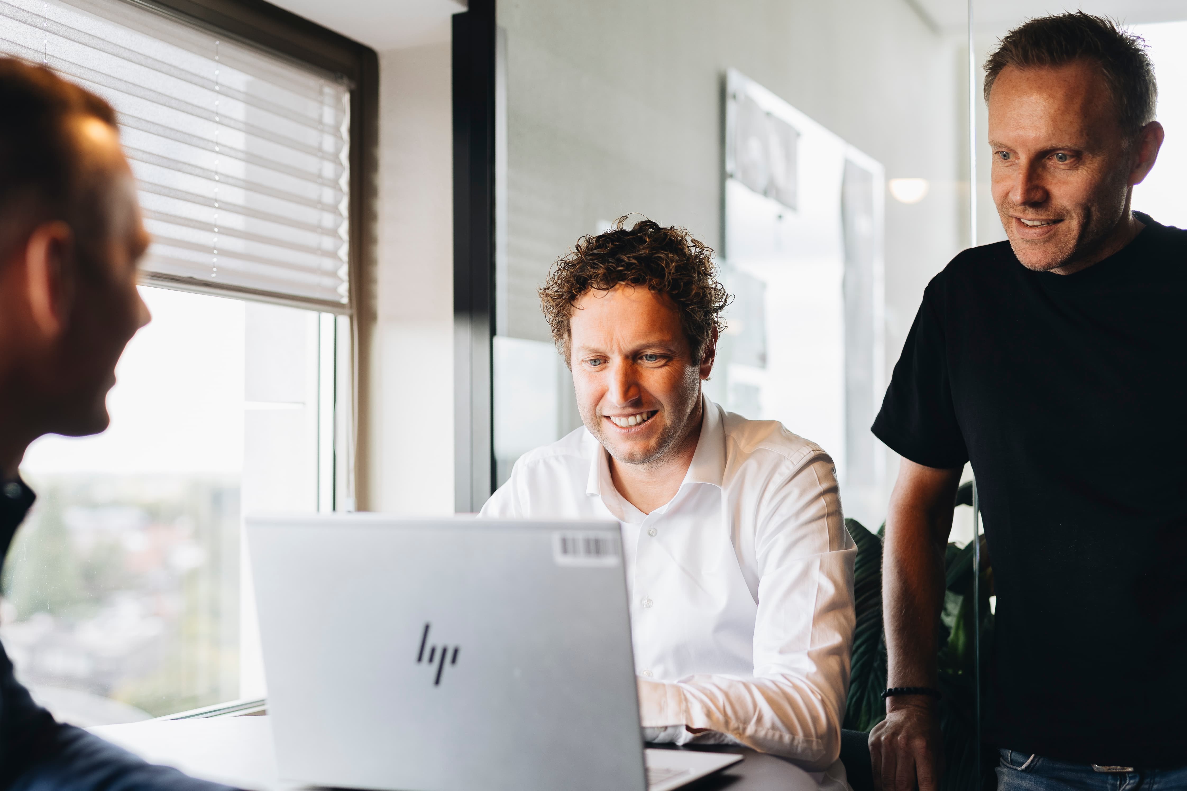Drie personen in een kantoor overleggen rond een laptop. Zakelijke teamvergadering met HP-laptop bij het raam