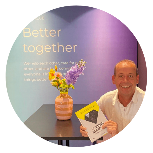 Man met wit overhemd houdt een boek vast naast een tafel met bloemen, voor een paarsblauwe muur met de tekst Better together