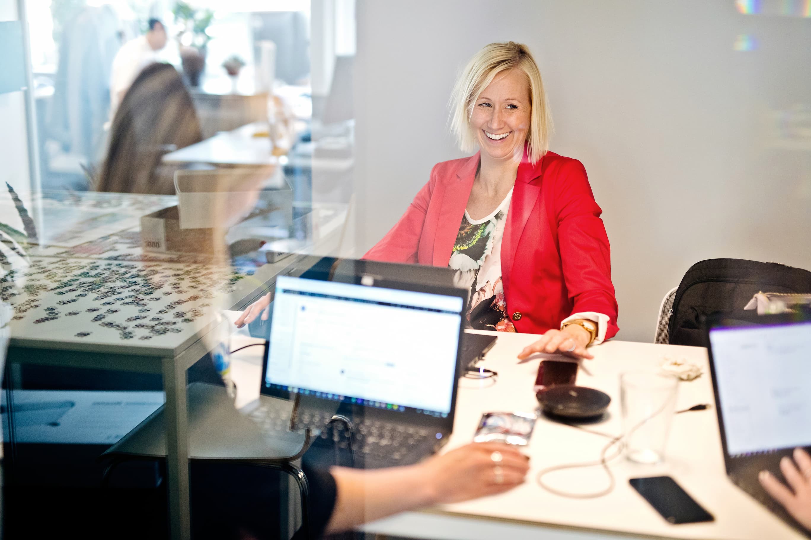 Kvinna i röd blazer sitter vid ett mötesbord i ett kontor med laptop och mobiltelefon. Avskild affärsmiljö med glasväggar och datorer