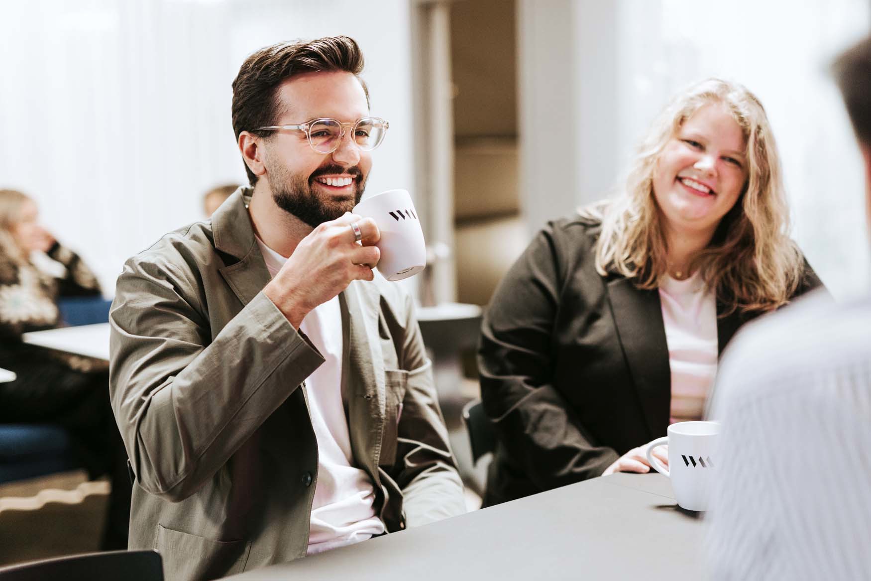 Två personer sitter vid ett bord och dricker kaffe ur vita muggar i en ljus konferensmiljö