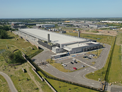 Stor industribygning med parkeringsplads og adgangsveje set fra luften. Lagerhal eller logistikcenter i åbent erhvervsområde
