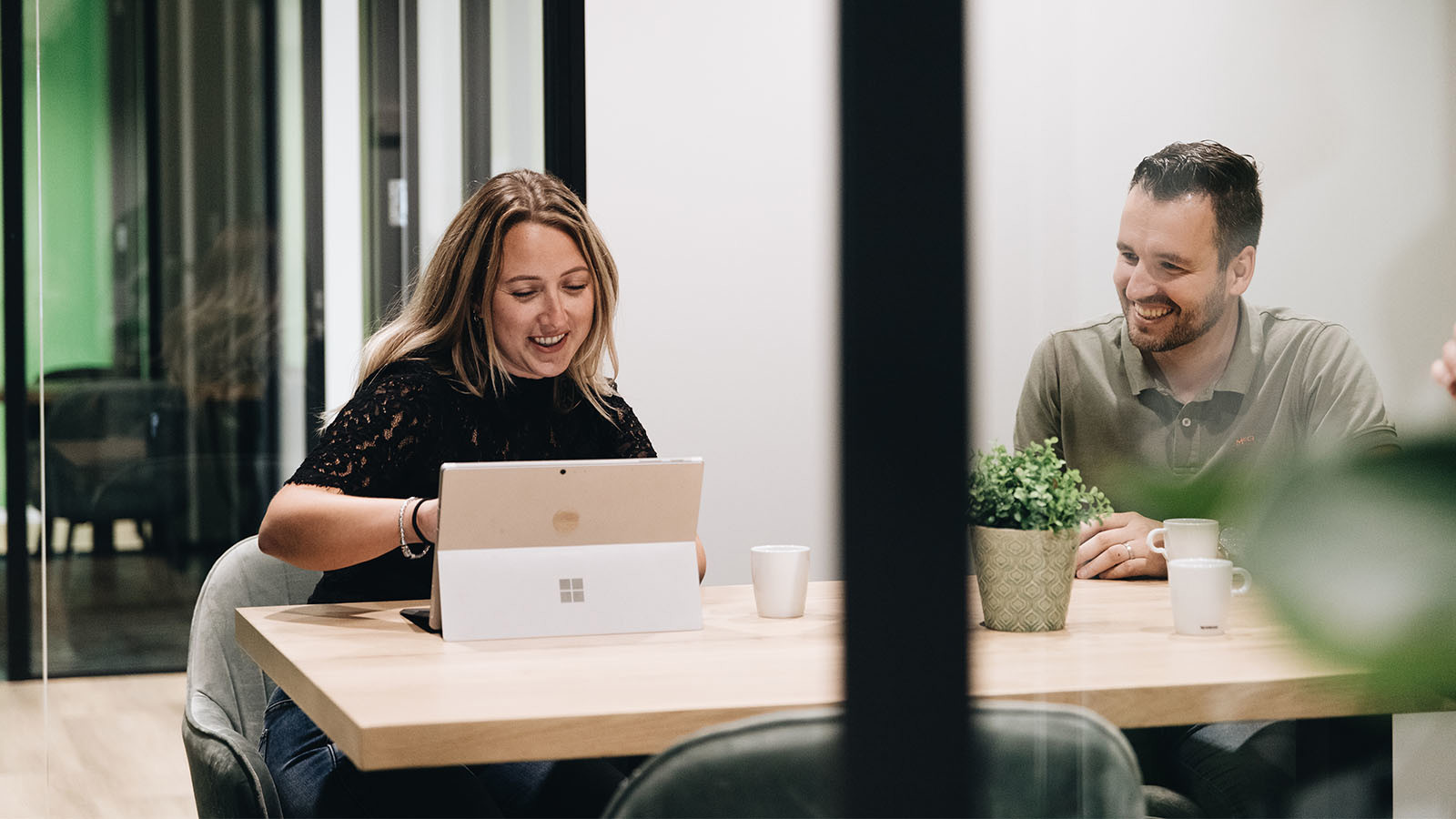 Två personer sitter vid ett mötesbord i ett modernt kontor med laptop, kaffe och växt