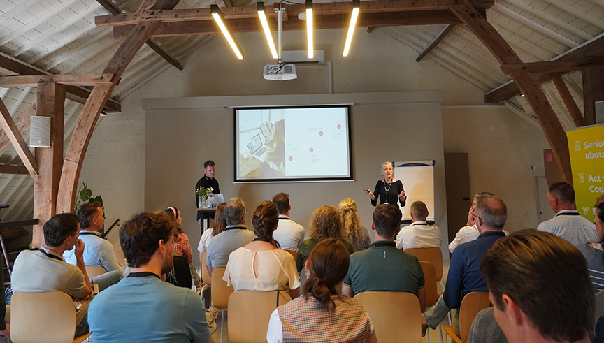 Zakelijke presentatie met spreker voor publiek in vergaderruimte, met projectiescherm en flip-over. Houten balken en moderne verlichting in een conferentieruimte