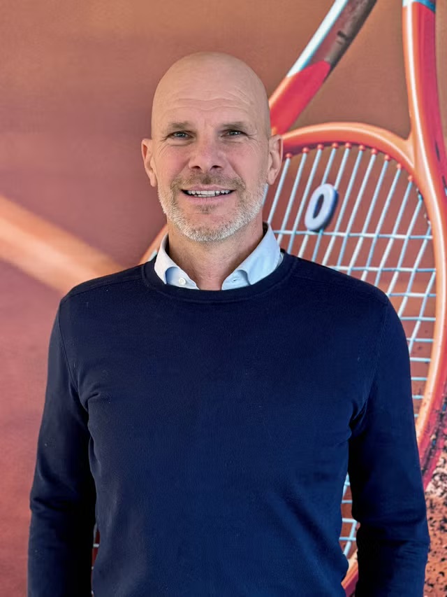 Man i mörkblå tröja framför en tennisracket i bakgrunden, med ansiktet maskerat
