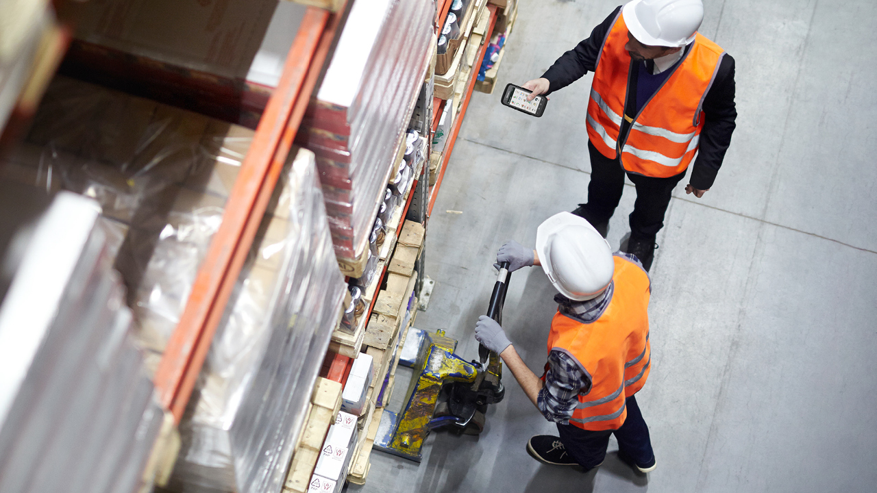 Två män i orange väst och vit arbetshatt står på ett lager med en handscanner