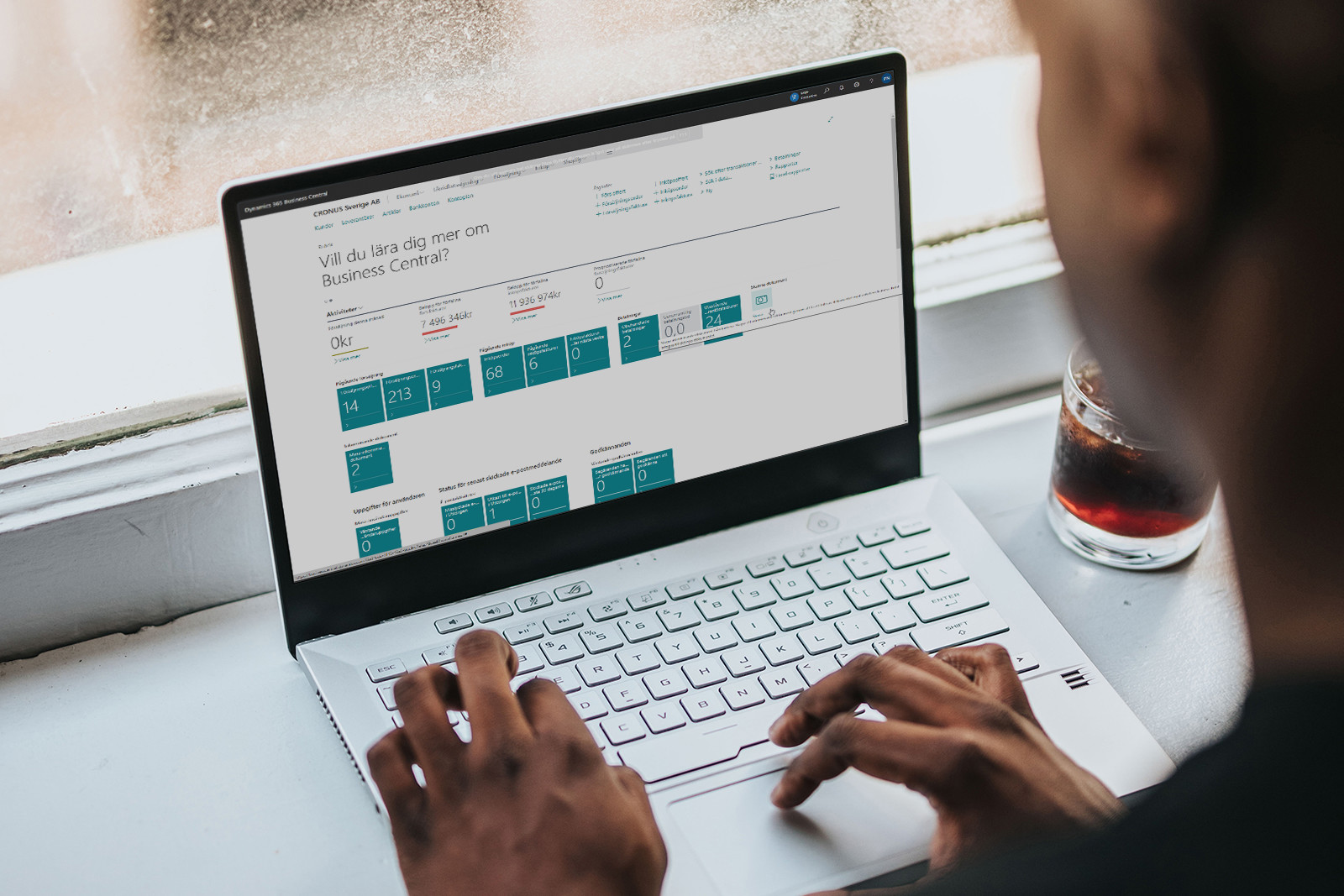 Person som skriver på en bärbar dator med Business Central-dashboard på skärmen och ett glas dryck bredvid