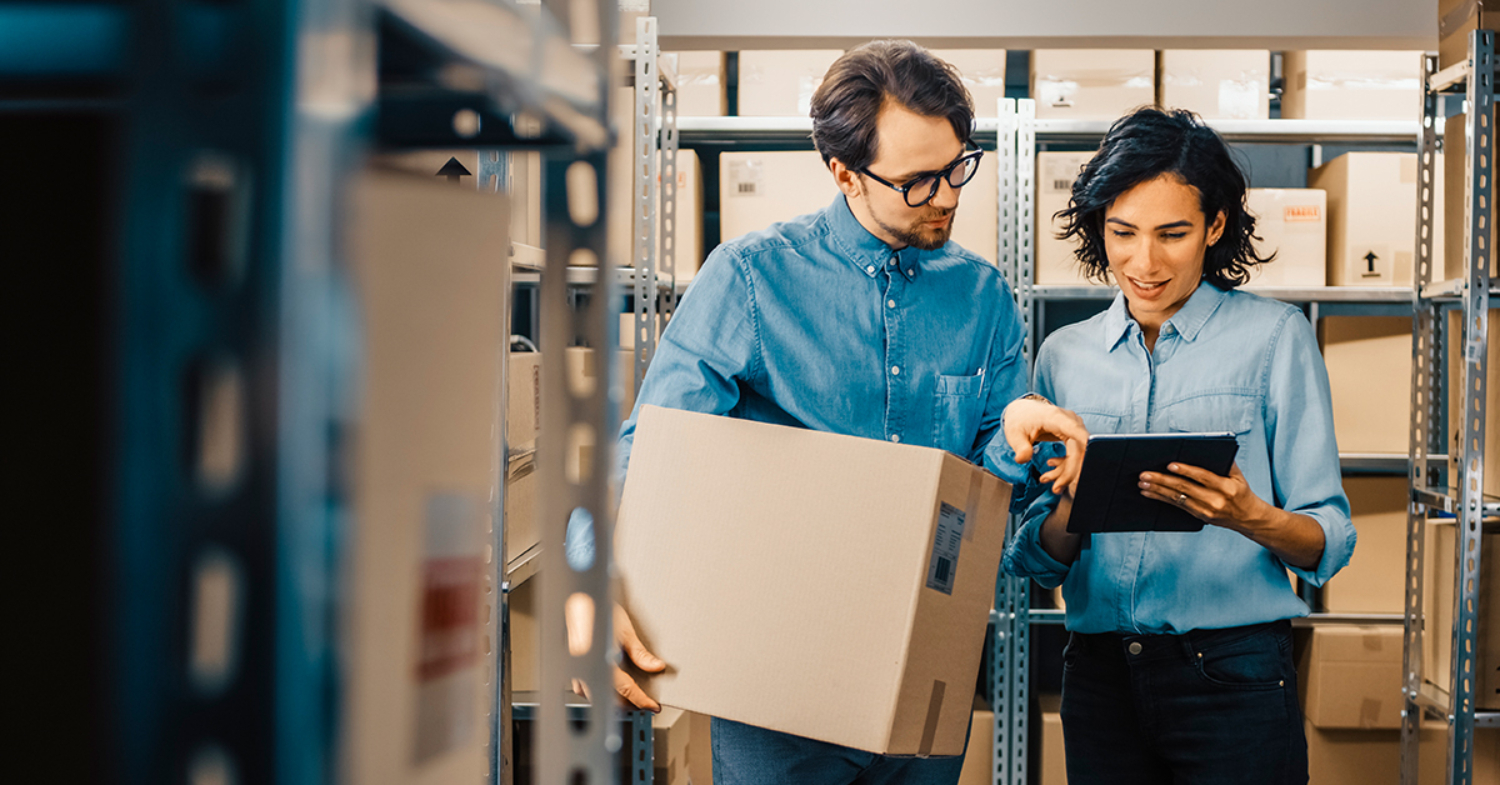 En man och en kvinna står bland lagerhyllor och tittar ihop på en ipad. 