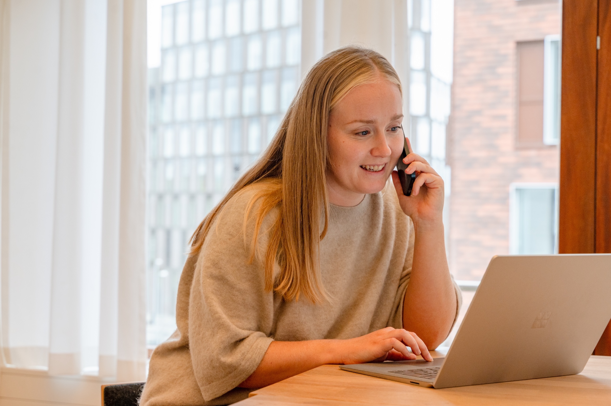 Woman talking on the phone, sitting at a desk in front of a laptop. A big window in the background.