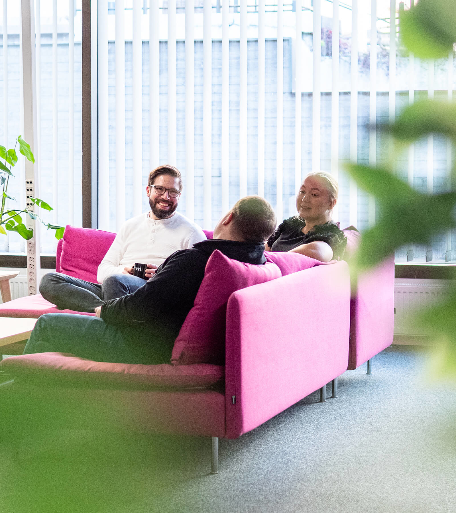 Tre personer sitter i en rosa soffa i ett ljust kontor eller loungeområde med stora fönster