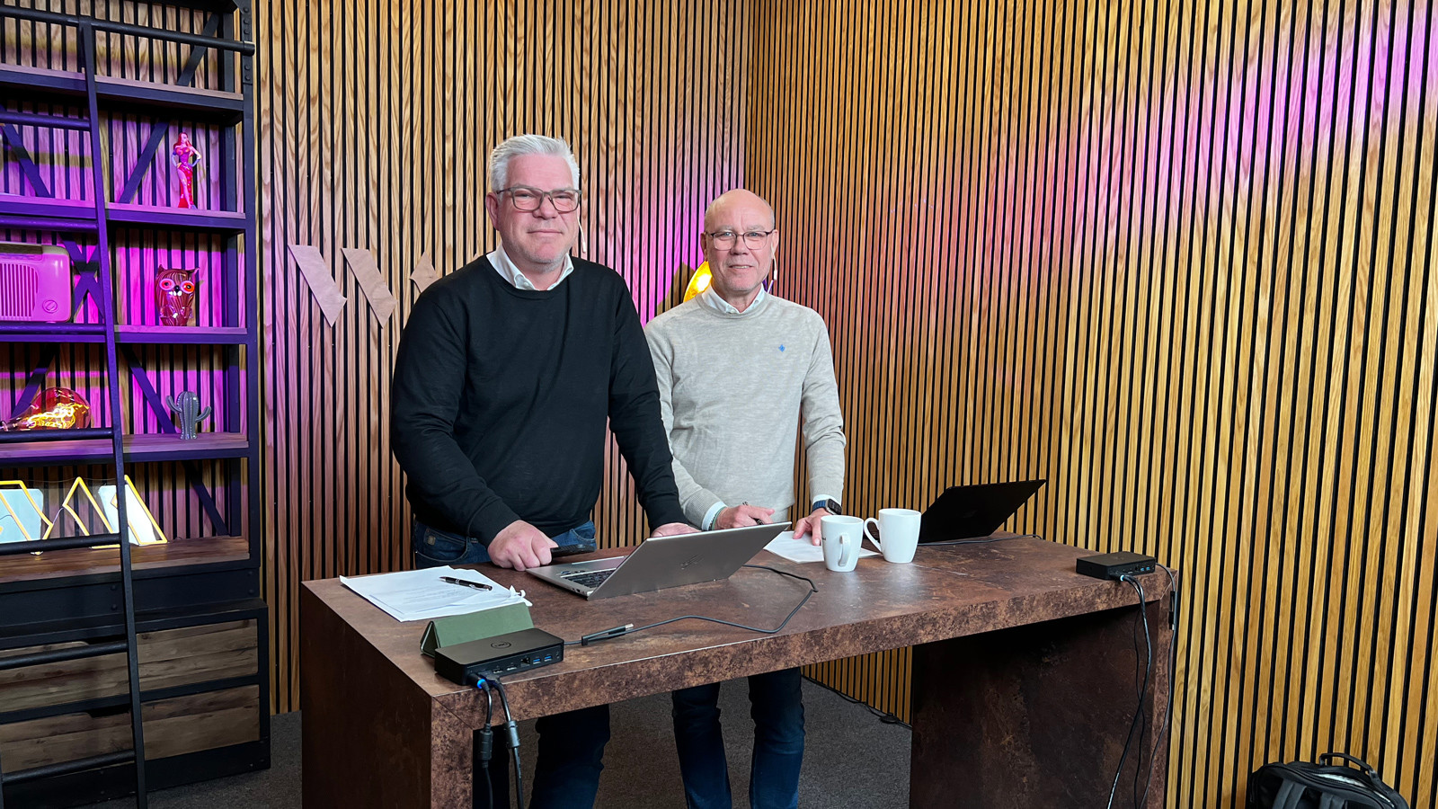Två personer står vid ett bord med laptops i ett kontorsliknande rum med träpaneler och färgstark belysning