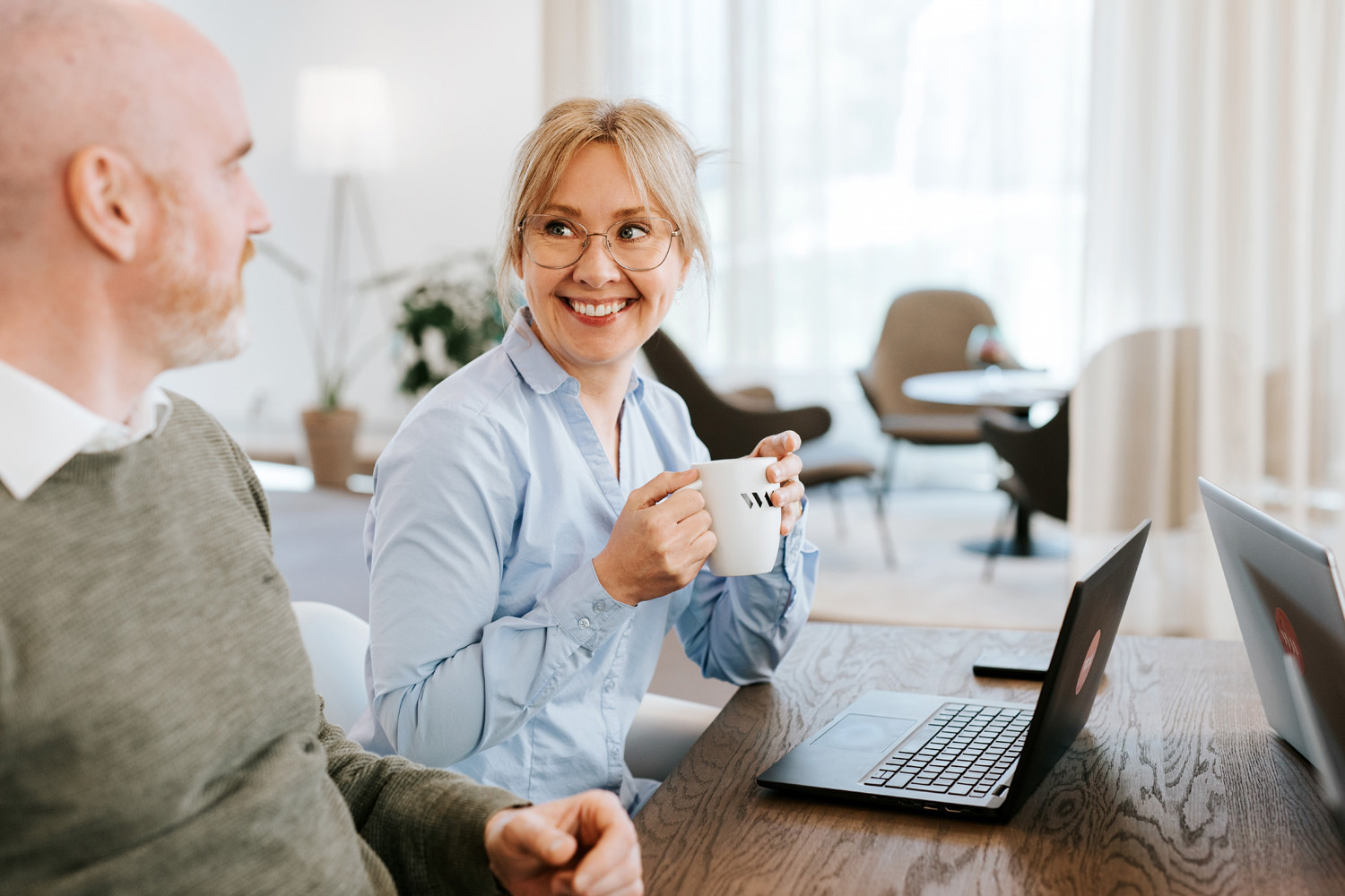 Två personer sitter vid ett bord med laptops i ett ljust kontor och samtalar över kaffe